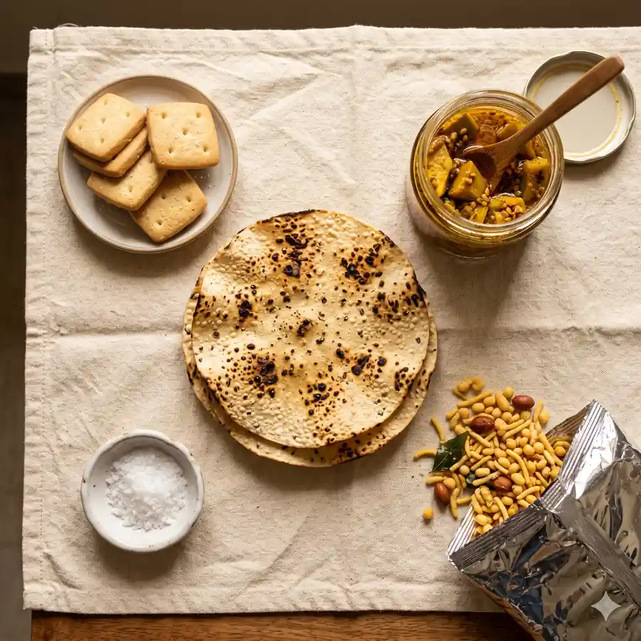 Roasted papad, mango achaar jar, namkeen packet, salted biscuits and a small dish of salt on a cream tablecloth.